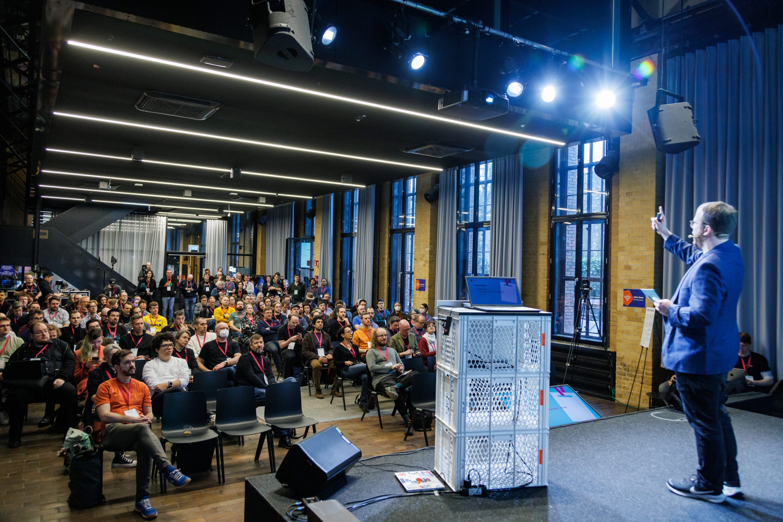 A picture from FOSS Backstage 2026. The photo was taken from behind the lectern overlooking the audience in front of the main stage. The room is filled with people listening to a man on stage. The stage lights create lens flair in the picture.